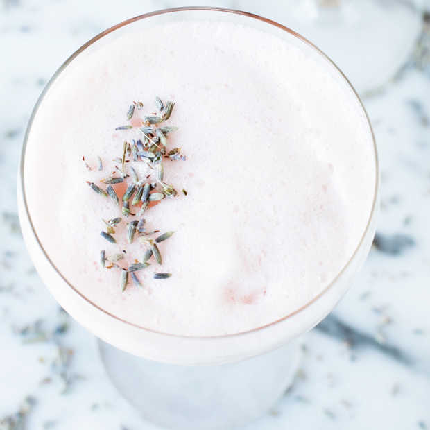 A frothy pink cocktail in a glass, garnished with dried lavender buds on top, sits on a light-colored marble surface.
