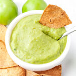 A bowl of green salsa dip with a tortilla chip partially dipped in it, surrounded by more chips and whole green tomatoes on a white surface.