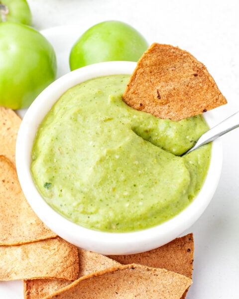 A bowl of green salsa dip with a tortilla chip partially dipped in it, surrounded by more chips and whole green tomatoes on a white surface.