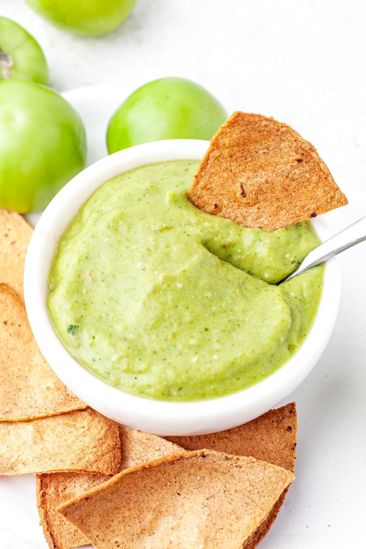 A bowl of green salsa dip with a tortilla chip partially dipped in it, surrounded by more chips and whole green tomatoes on a white surface.