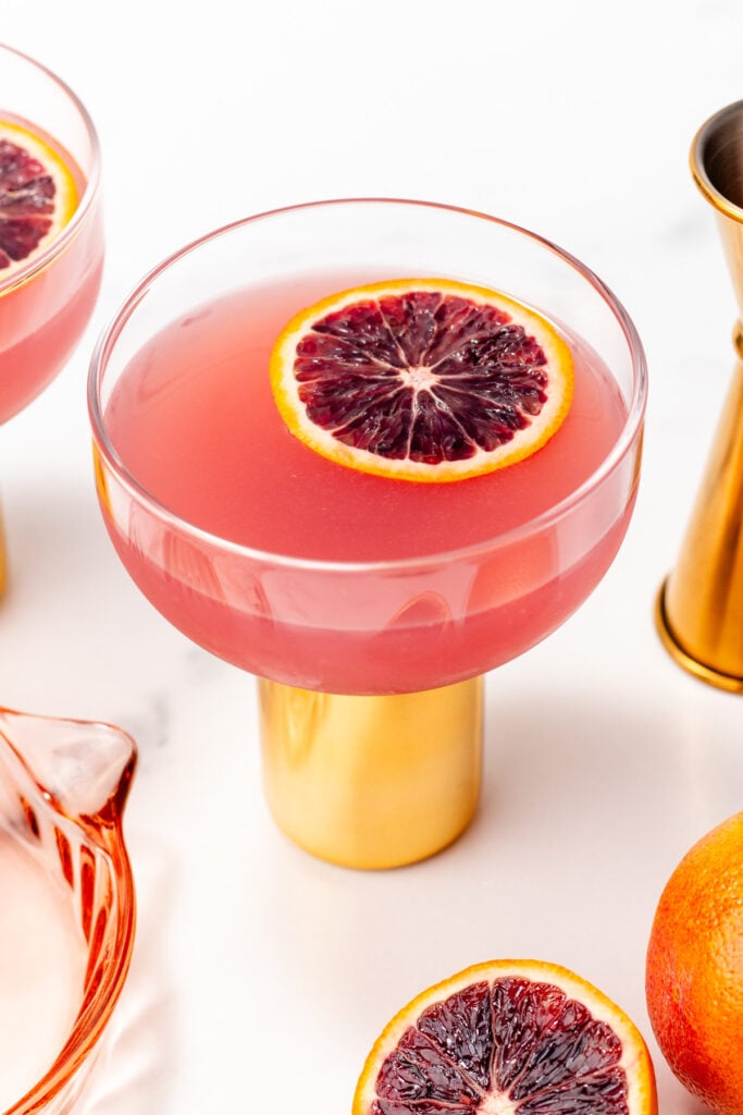 A pink cocktail in a gold-based glass, garnished with a slice of blood orange, sits on a white surface near a jigger and blood orange slices.