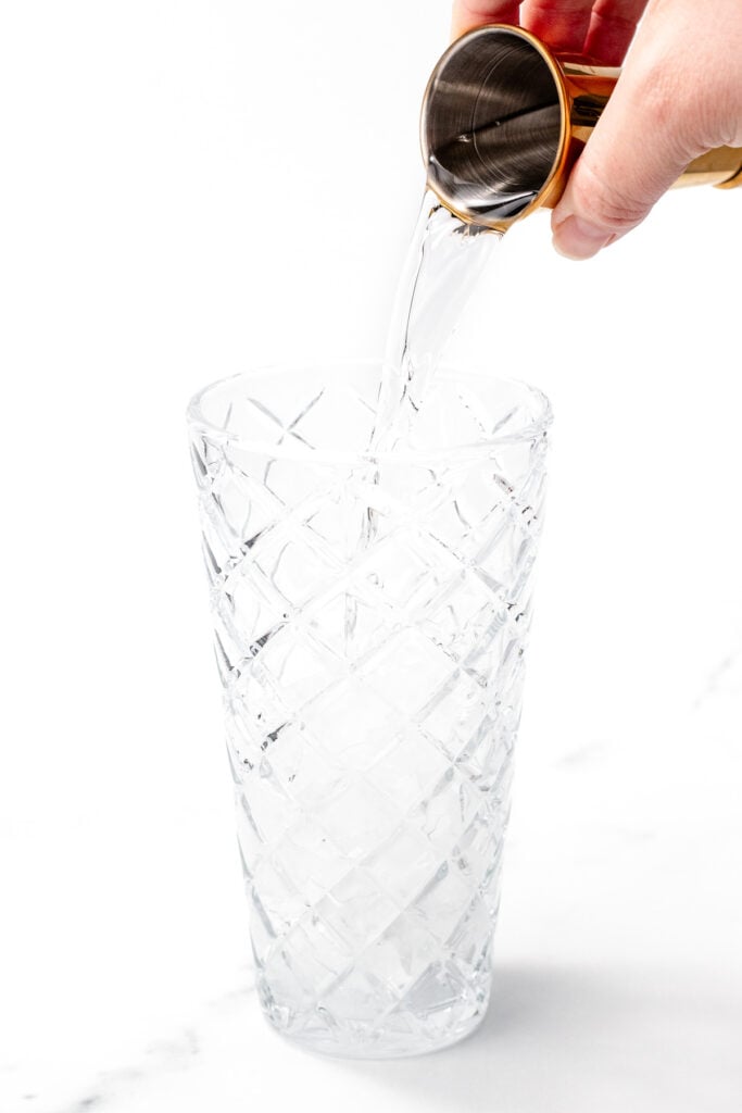 A hand pours clear liquid from a metal jigger into a tall, empty, patterned glass on a white surface.