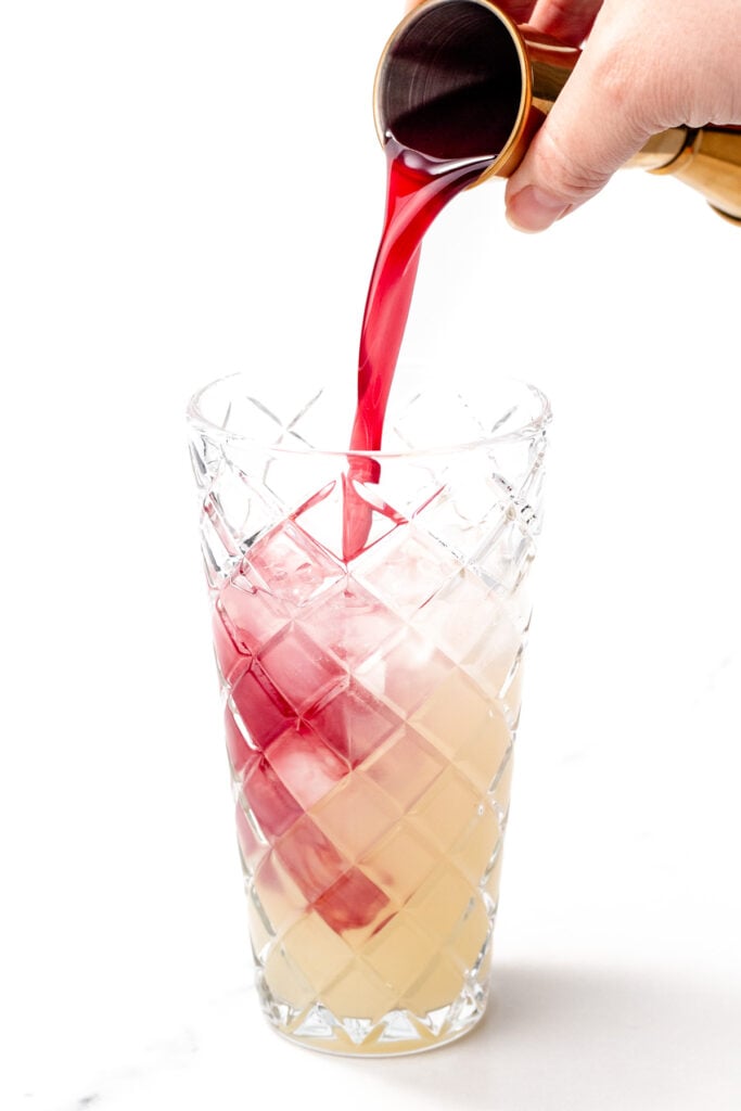 A hand pours red liquid from a jigger into a tall glass of ice and pale yellow liquid.