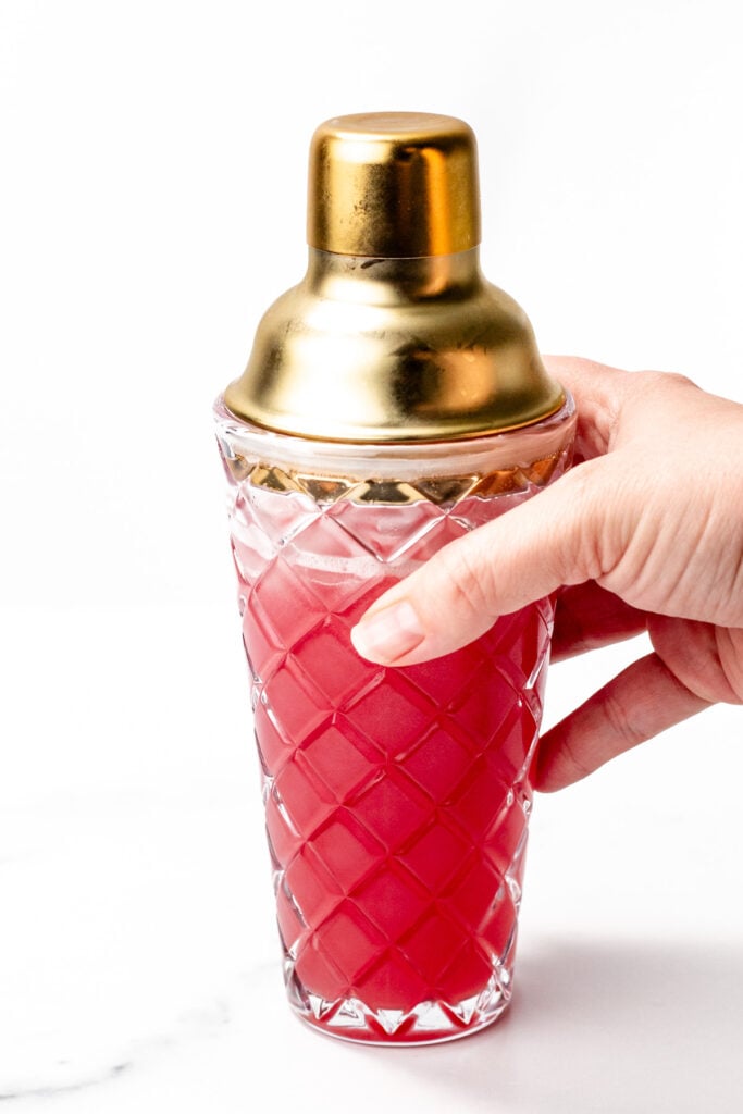 A hand holding a glass cocktail shaker with a gold lid, filled with a red liquid, against a white background.