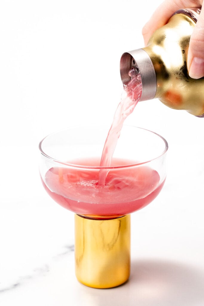 A pink cocktail is being poured from a gold cocktail shaker into a glass with a gold base on a white background.