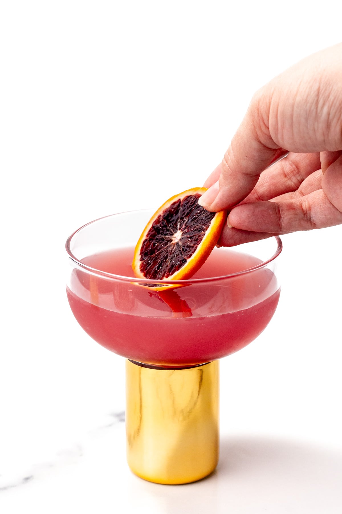 A hand places a slice of blood orange into a pink cocktail in a glass with a gold base, set against a white background.