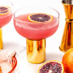 A glass with a gold stem contains a pink cocktail garnished with a blood orange slice; nearby are whole and halved blood oranges, a measuring jigger, and a glass citrus juicer.