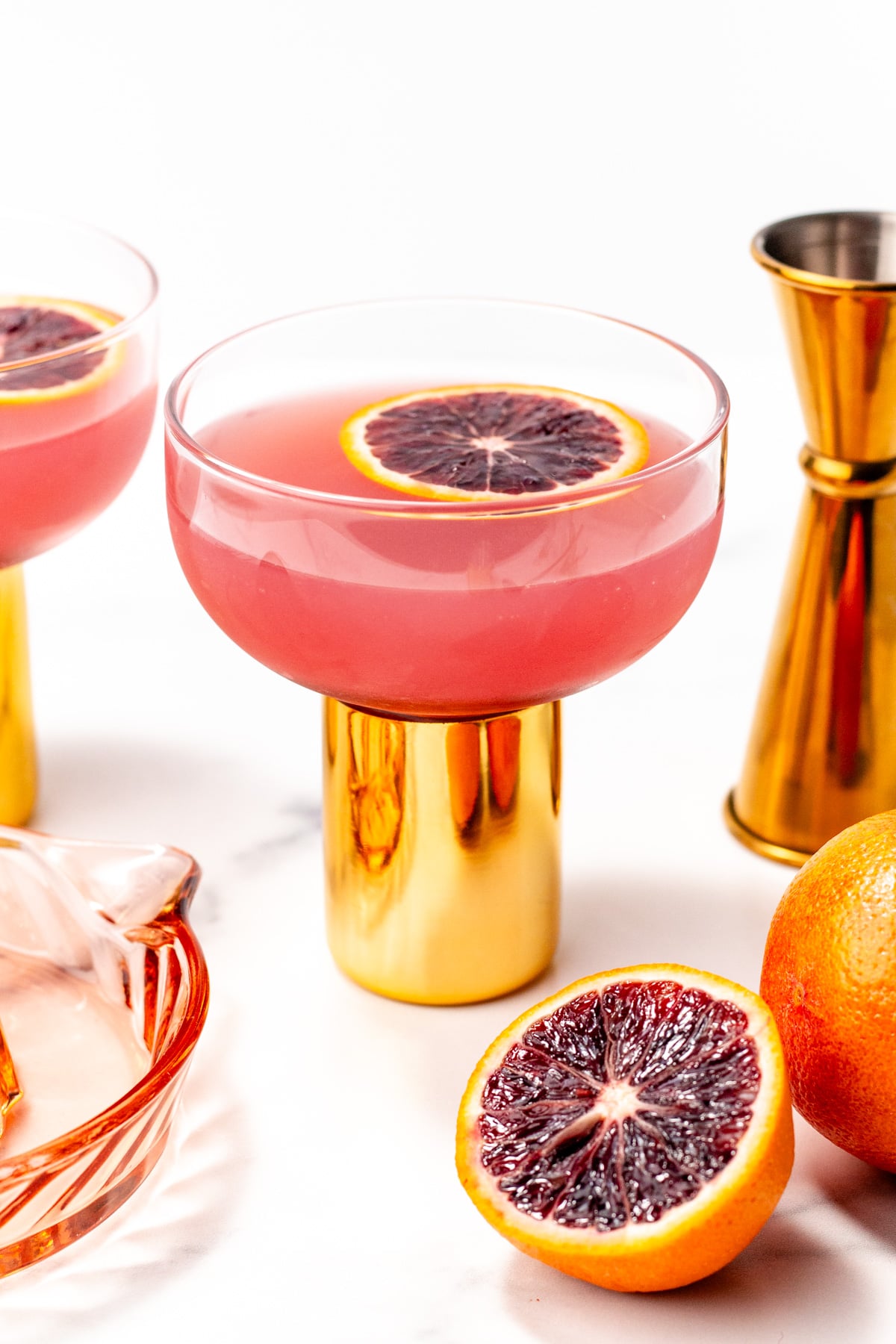 A glass with a gold stem contains a pink cocktail garnished with a blood orange slice; nearby are whole and halved blood oranges, a measuring jigger, and a glass citrus juicer.