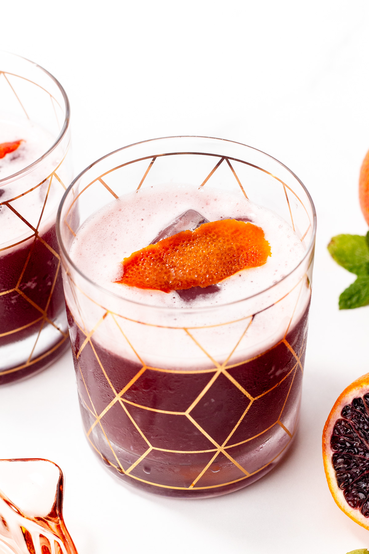 A glass with geometric gold pattern filled with a dark red frothy drink, ice, and a citrus peel garnish on top. Another glass and blood orange slices are partially visible nearby.