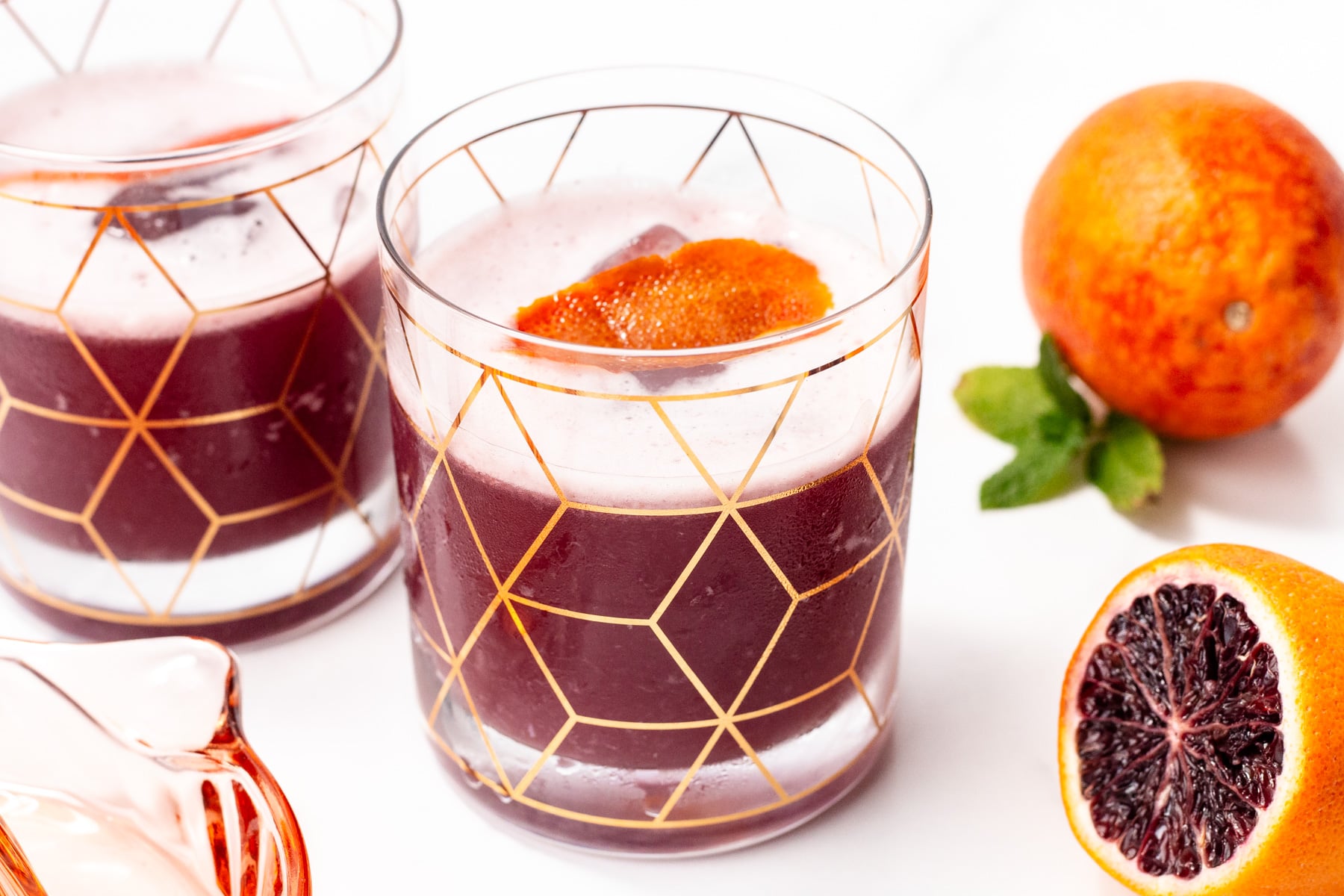 Two glasses of dark red juice with orange peel garnish, next to a whole blood orange and a halved blood orange on a white surface.
