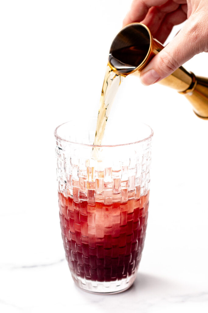 A hand pours a golden liquid from a jigger into a glass with a red beverage and ice, against a white background.