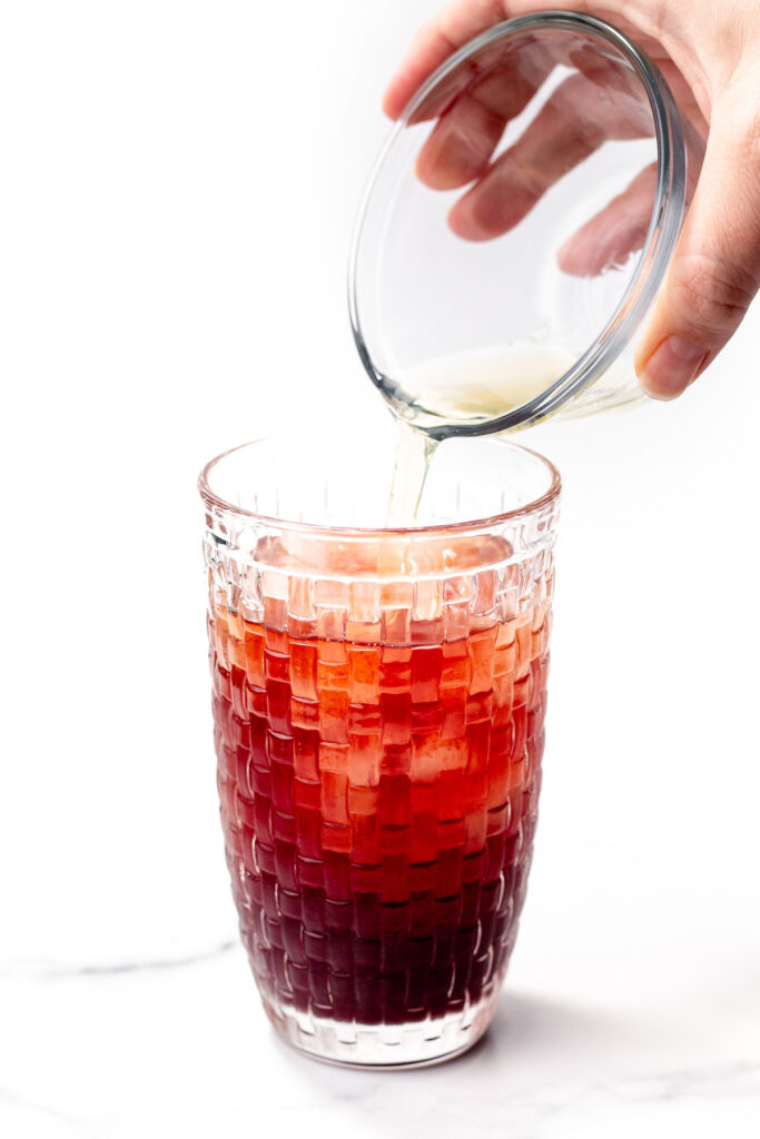 A hand pours a light-colored liquid from a small glass bowl into a tall glass filled with a dark red beverage.