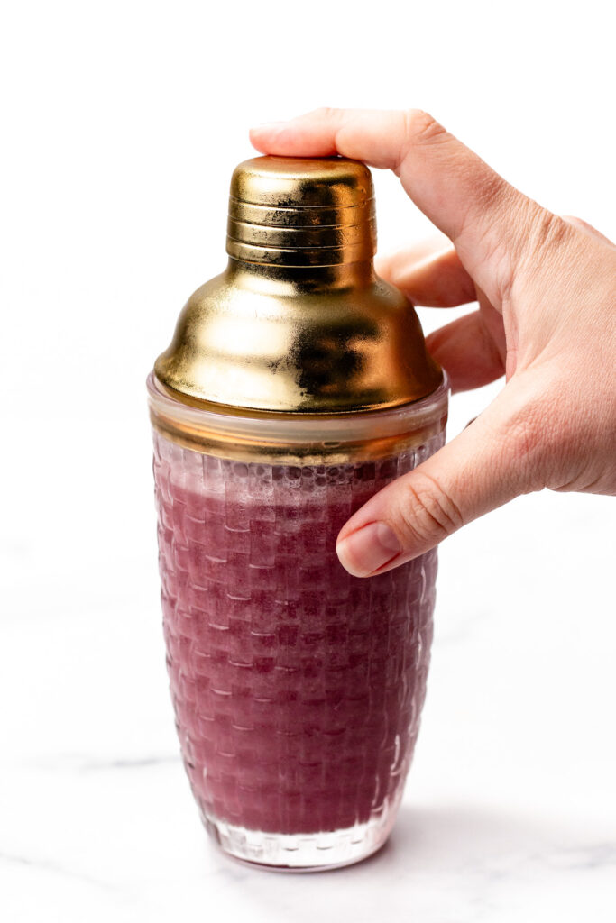A hand holds a textured glass cocktail shaker with a gold-colored lid, filled with a purple liquid, against a white background.