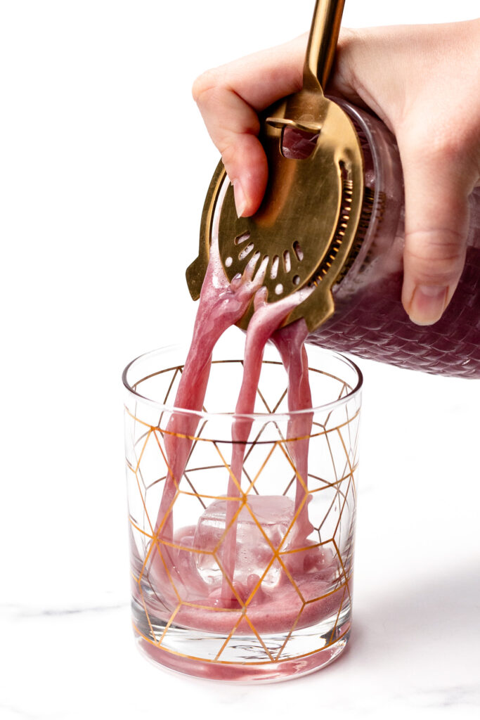 A hand pours a pink beverage through a gold cocktail strainer into a glass with ice and a geometric gold pattern.