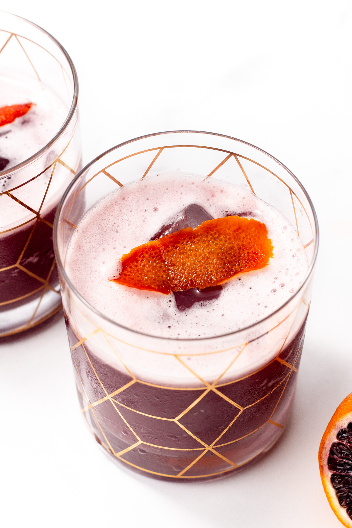A glass of dark red cocktail with a frothy top, garnished with an orange peel, sits on a white surface. Another glass and a sliced orange are partially visible nearby.