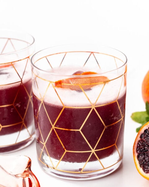 A glass with a geometric gold pattern holds a dark red beverage, garnished with a citrus slice. Another similar glass and blood oranges are partially visible nearby.