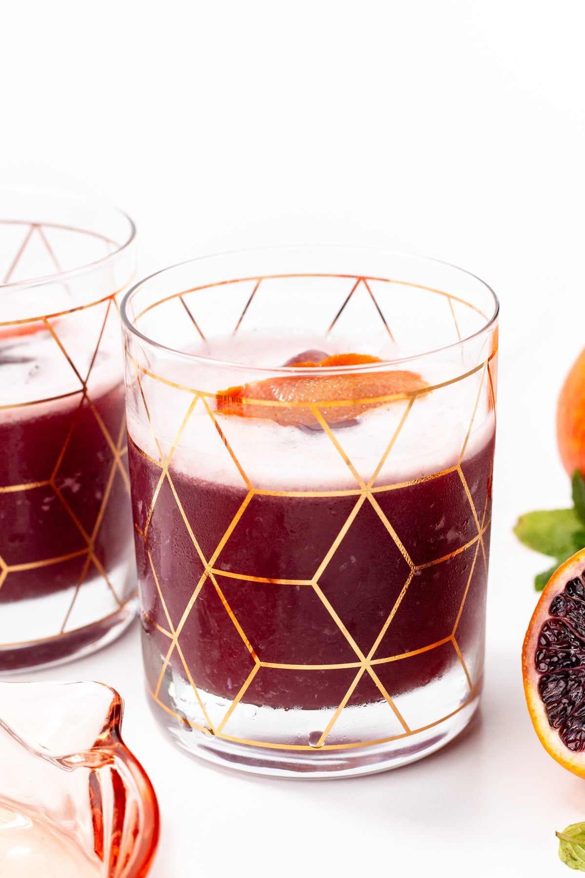 A glass with a geometric gold pattern holds a dark red beverage, garnished with a citrus slice. Another similar glass and blood oranges are partially visible nearby.