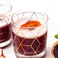 A glass with a geometric gold pattern filled with a dark purple-red drink and a slice of orange garnish, placed on a white surface.
