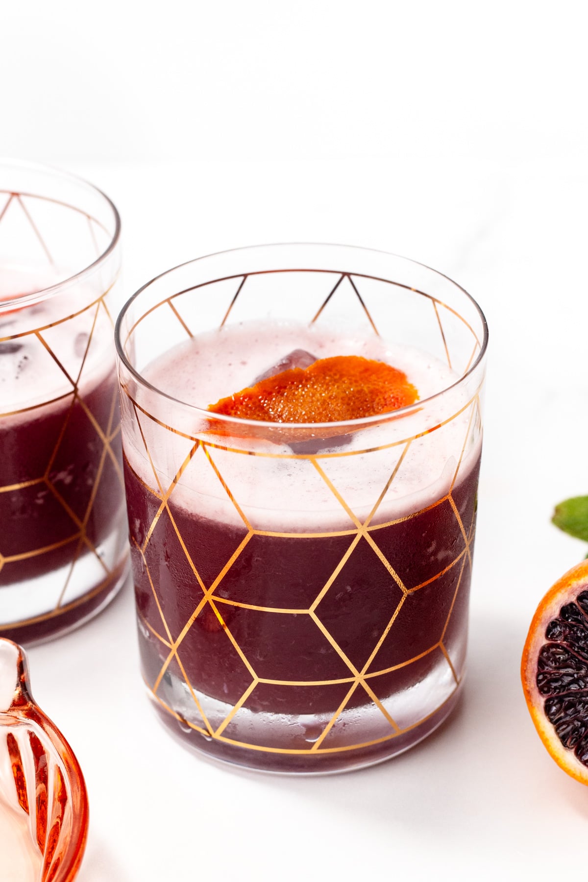 A glass with a geometric gold pattern filled with a dark purple-red drink and a slice of orange garnish, placed on a white surface.