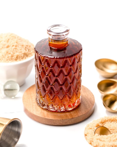 A decorative glass bottle filled with dark liquid sits on a wooden coaster, surrounded by bowls of brown sugar and gold measuring spoons on a white background.
