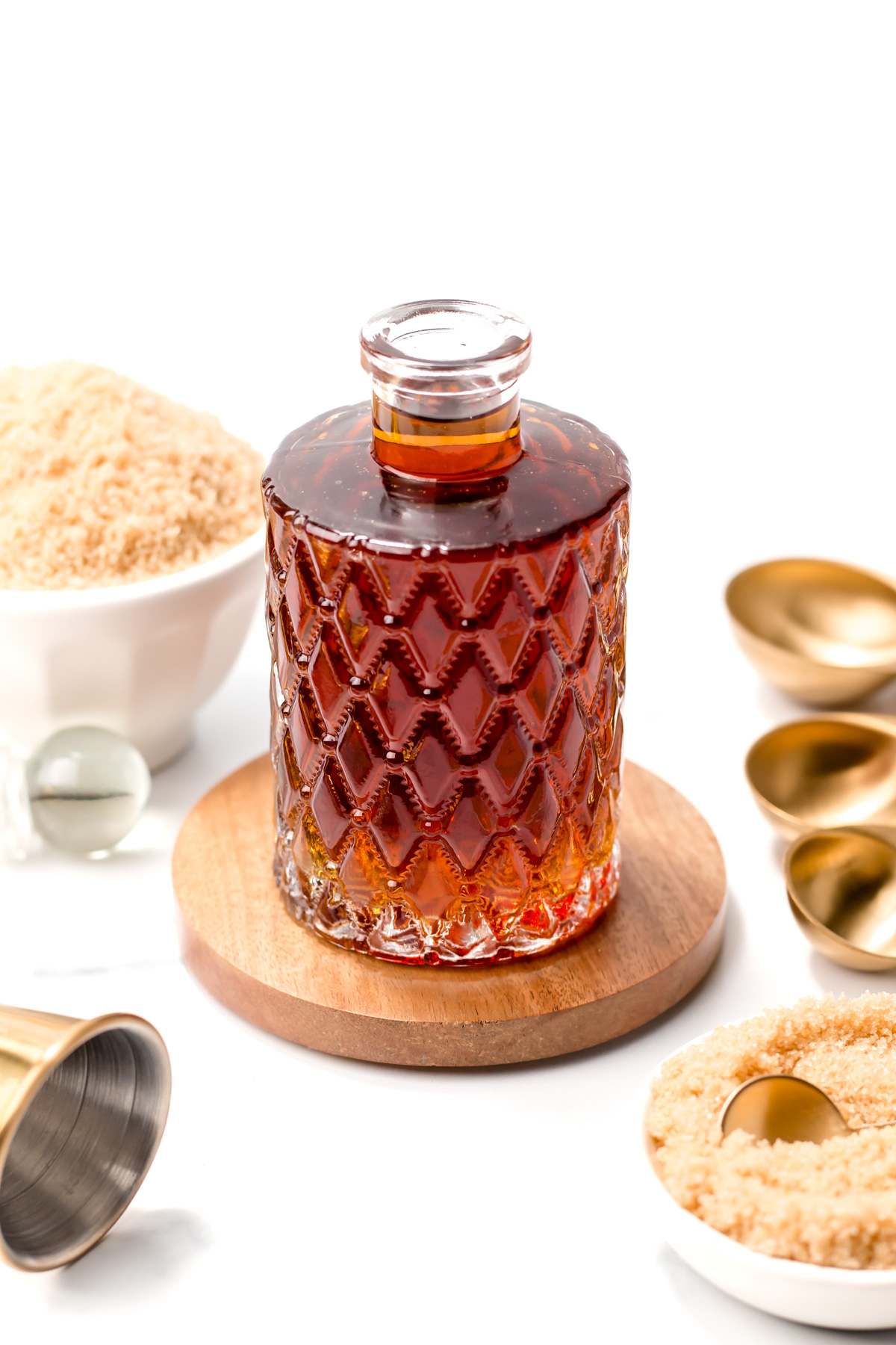 A decorative glass bottle filled with dark liquid sits on a wooden coaster, surrounded by bowls of brown sugar and gold measuring spoons on a white background.