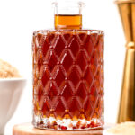 A clear glass decanter with diamond pattern contains a dark amber liquid, placed on a wooden coaster, with bowls of brown sugar and a gold jigger nearby.