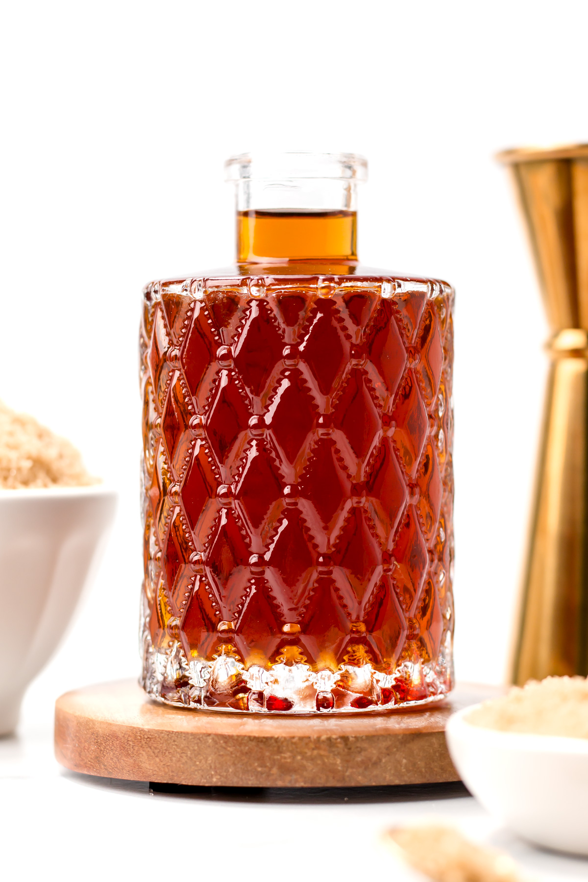 A clear glass decanter with diamond pattern contains a dark amber liquid, placed on a wooden coaster, with bowls of brown sugar and a gold jigger nearby.