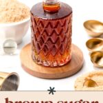 A glass bottle of brown sugar syrup sits on a wooden coaster, surrounded by gold measuring spoons and a bowl of brown sugar.