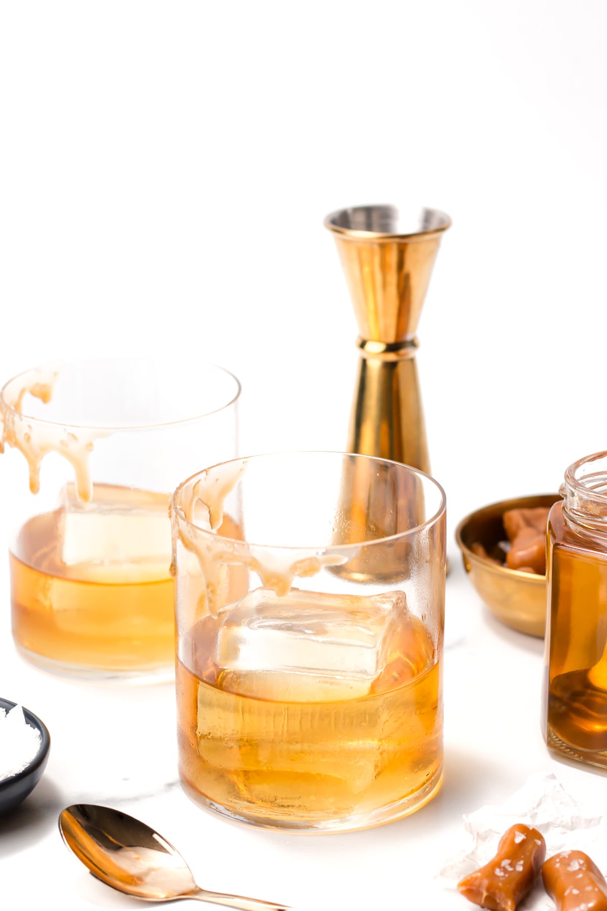 Two glasses of caramel whiskey on ice sit on a white surface near a gold jigger, a spoon, caramel candies, and a jar of caramel sauce.