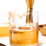 A glass of whiskey with a large ice cube sits on a white surface, with liquid spilled down the side. Gold barware and a spoon are in the background.