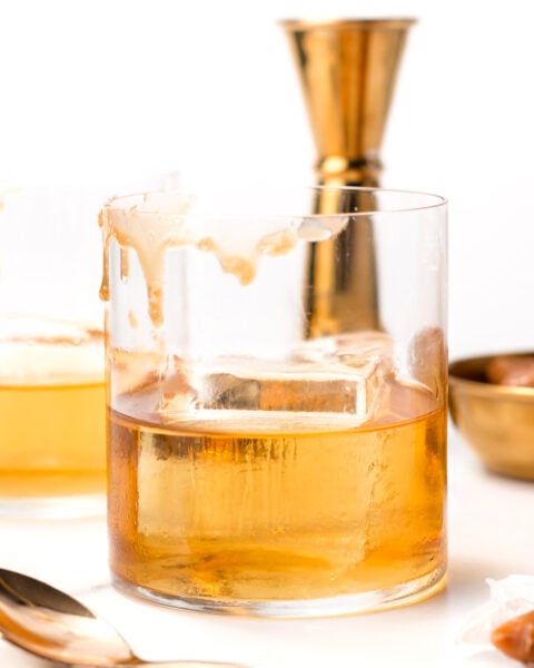 A glass of whiskey with a large ice cube sits on a white surface, with liquid spilled down the side. Gold barware and a spoon are in the background.