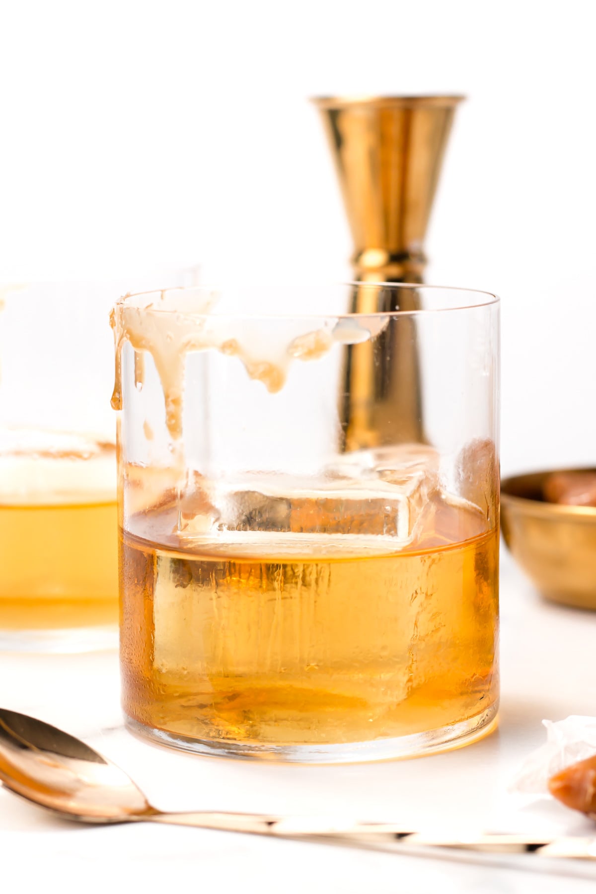 A glass of whiskey with a large ice cube sits on a white surface, with liquid spilled down the side. Gold barware and a spoon are in the background.