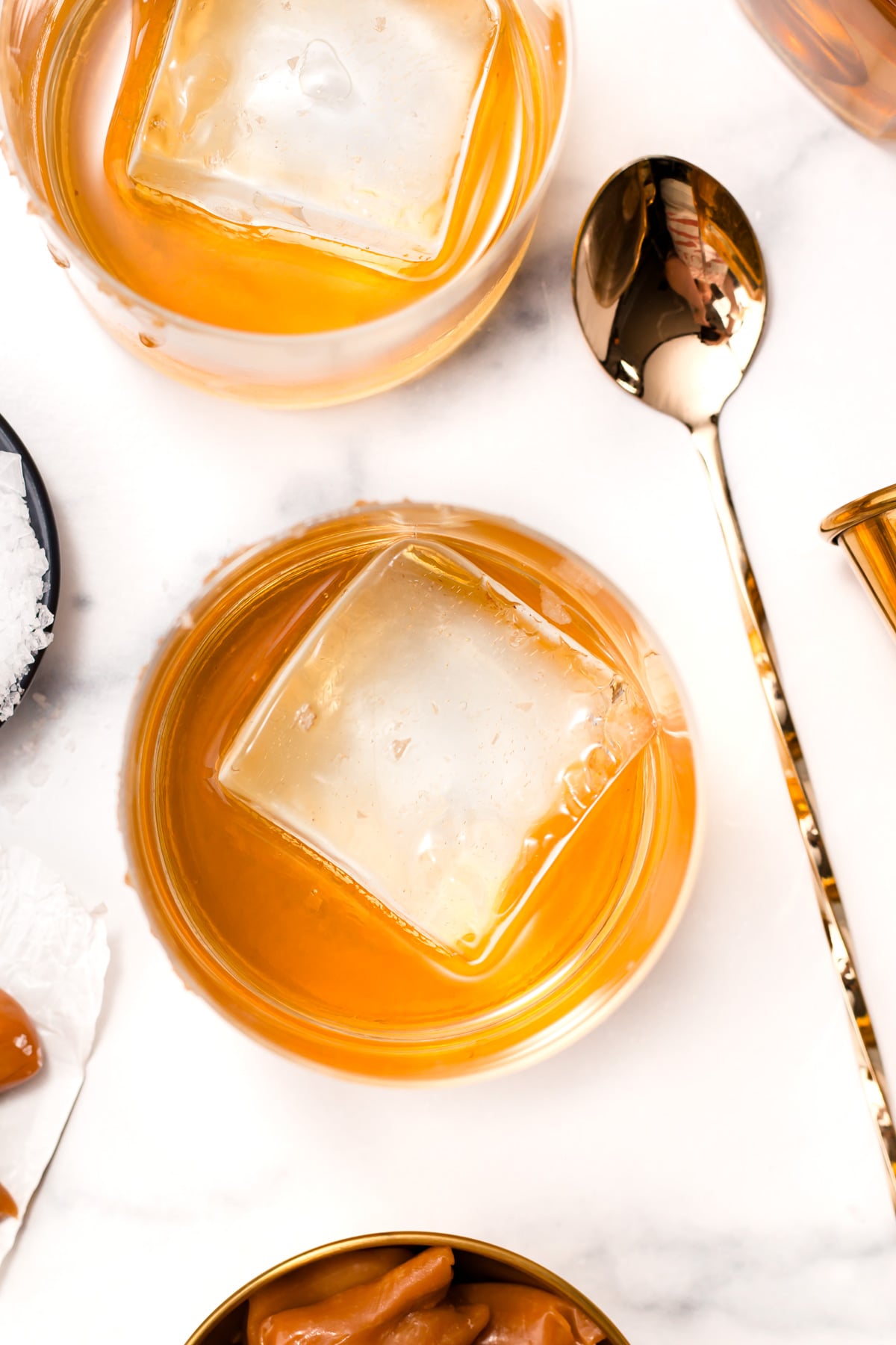 Two glasses of amber-colored liquor with large square ice cubes on a white surface, next to a gold spoon and caramel candies.