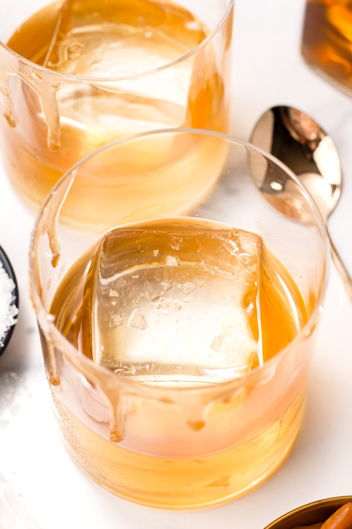 Two glasses of whiskey with large square ice cubes on a white surface, next to a metal spoon.