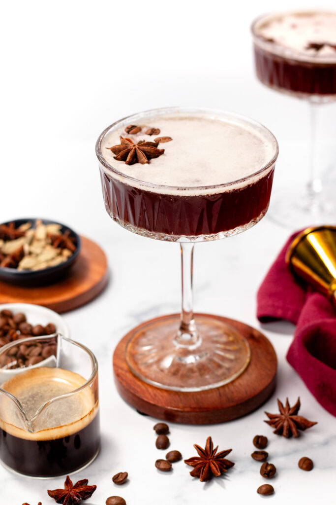 A coupe glass filled with a dark espresso martini, garnished with star anise, sits on a coaster surrounded by coffee beans, star anise, and a small glass of espresso.