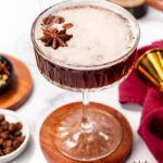 A cocktail served in a coupe glass, garnished with whole star anise, with coffee beans and cocktail tools in the background.