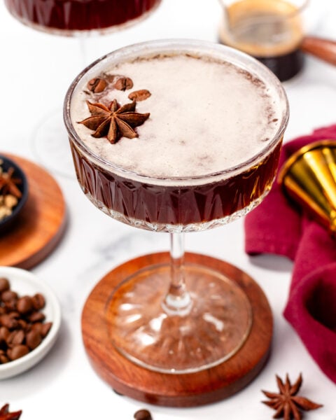 A cocktail served in a coupe glass, garnished with whole star anise, with coffee beans and cocktail tools in the background.