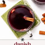 A glass mug of Danish mulled wine garnished with cinnamon sticks is placed on a green napkin, with additional cinnamon sticks and a bowl in the background.