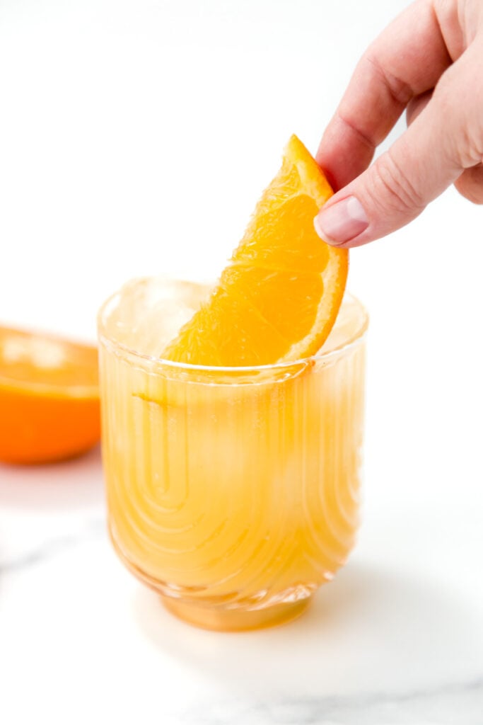 A hand places an orange slice garnish into a glass of iced orange-colored beverage on a white surface.