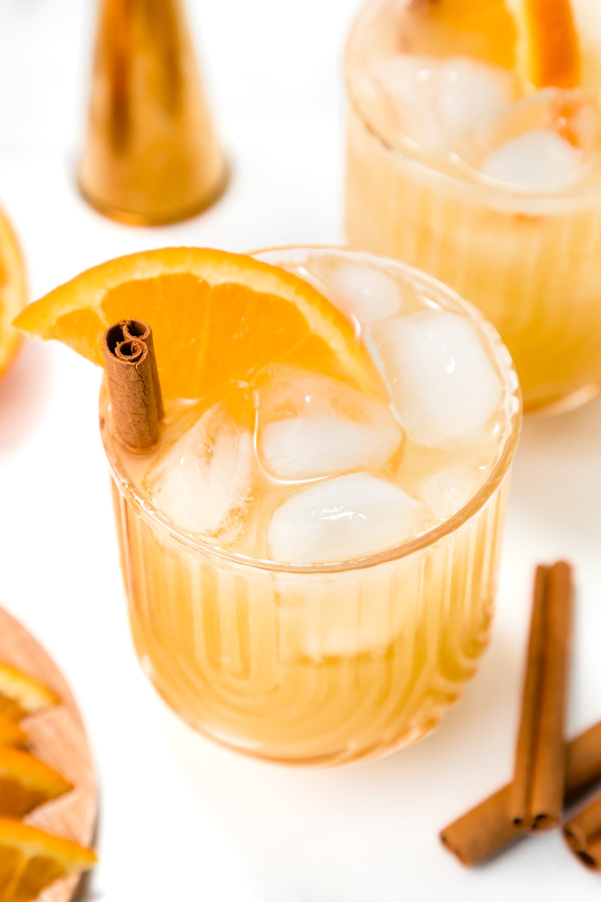 A glass filled with an iced orange-colored drink, garnished with an orange slice and a cinnamon stick. Another similar glass and cinnamon sticks are visible in the background.