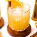 A glass of iced orange-colored drink garnished with an orange slice and a cinnamon stick, placed on a wooden coaster with cocktail tools and orange slices nearby.