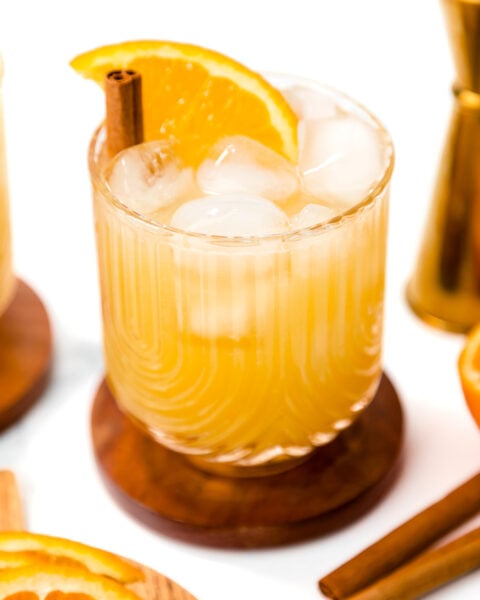 A glass of iced orange-colored drink garnished with an orange slice and a cinnamon stick, placed on a wooden coaster with cocktail tools and orange slices nearby.