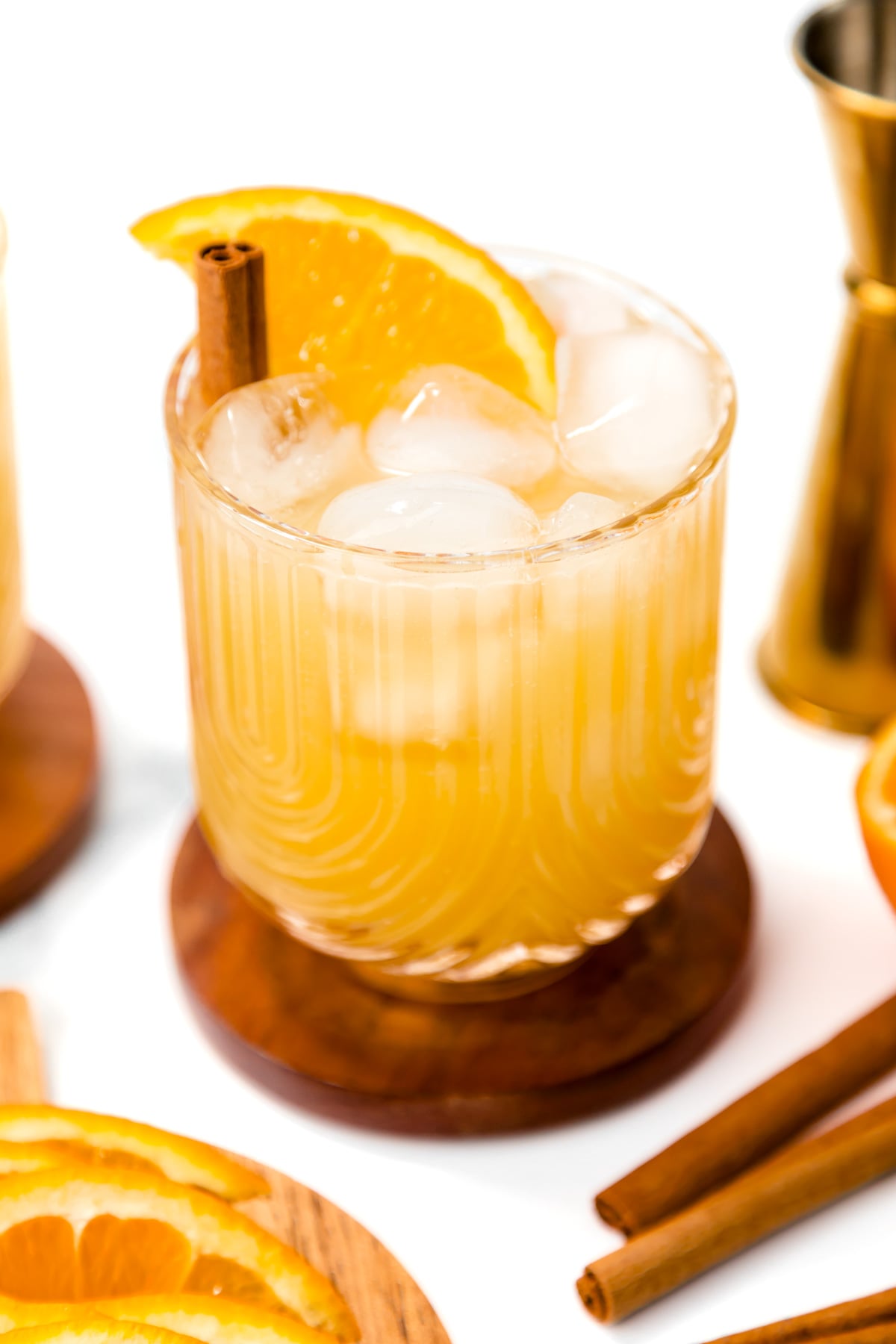 A glass of iced orange-colored drink garnished with an orange slice and a cinnamon stick, placed on a wooden coaster with cocktail tools and orange slices nearby.