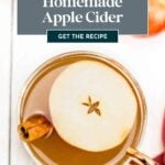 Top-down view of a glass mug filled with apple cider, garnished with a round apple slice and a cinnamon stick, with text overlay promoting a homemade apple cider recipe.