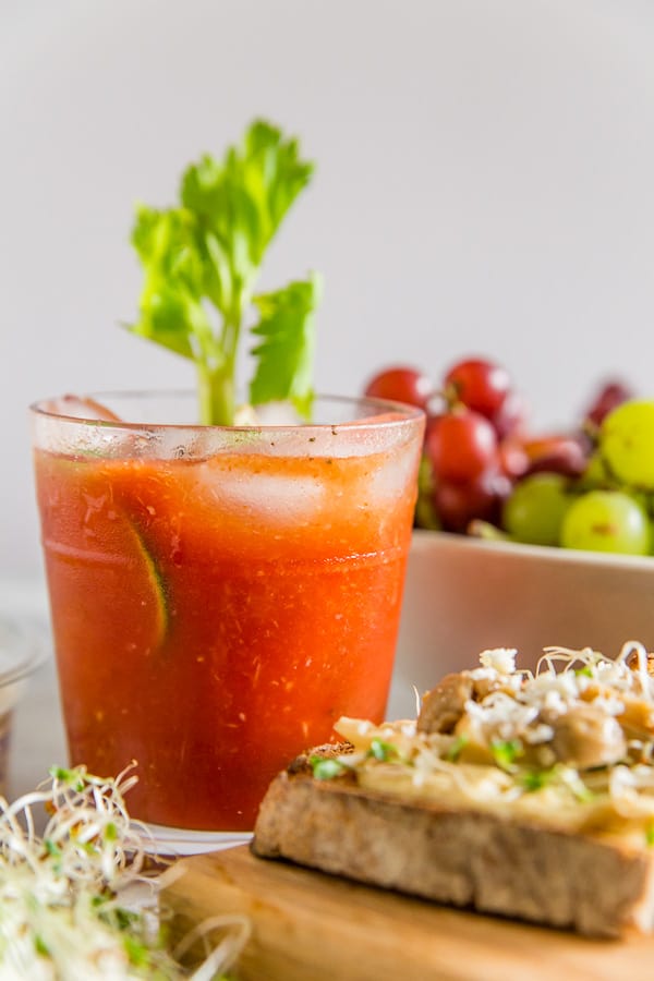 A glass of tomato juice with ice and celery, a slice of toast with toppings, and a bowl of grapes in the background.