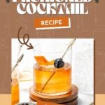 A glass of Old Fashioned cocktail garnished with an orange peel and a cherry, set on a wooden coaster with cocktail-making tools nearby.