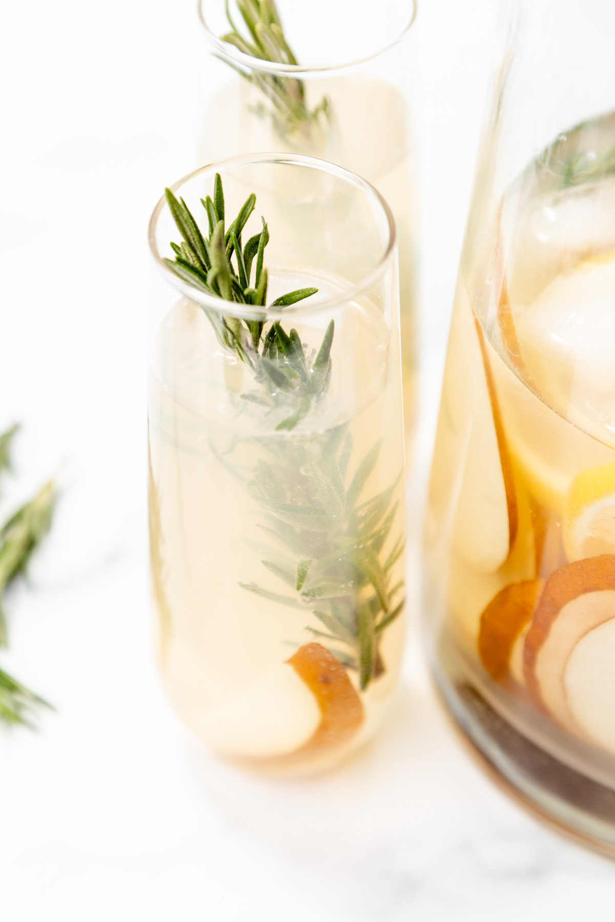 Two glasses and a pitcher of light-colored drink with ice, pear slices, and rosemary sprigs, set on a white surface.