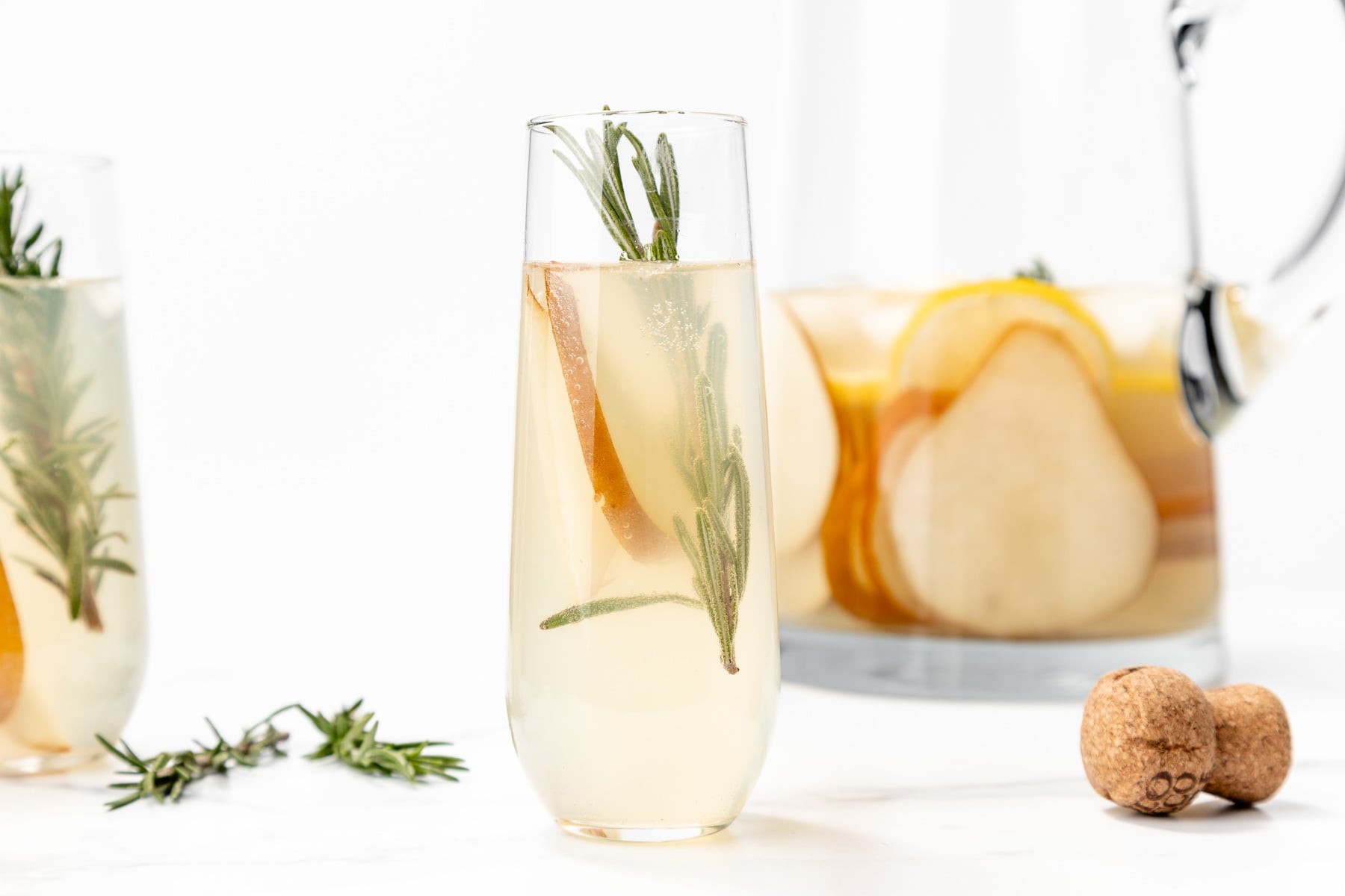 A glass of pale yellow beverage garnished with rosemary and a pear slice, with a pitcher of similar drink and a cork in the background.
