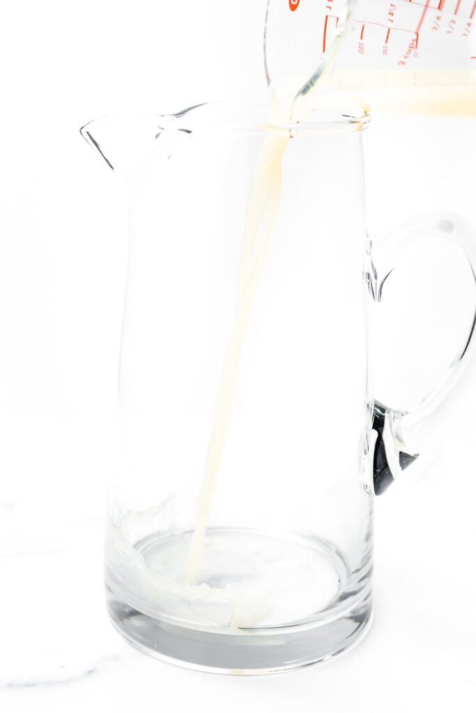 Milk is being poured from a measuring cup into a clear glass pitcher on a white background.