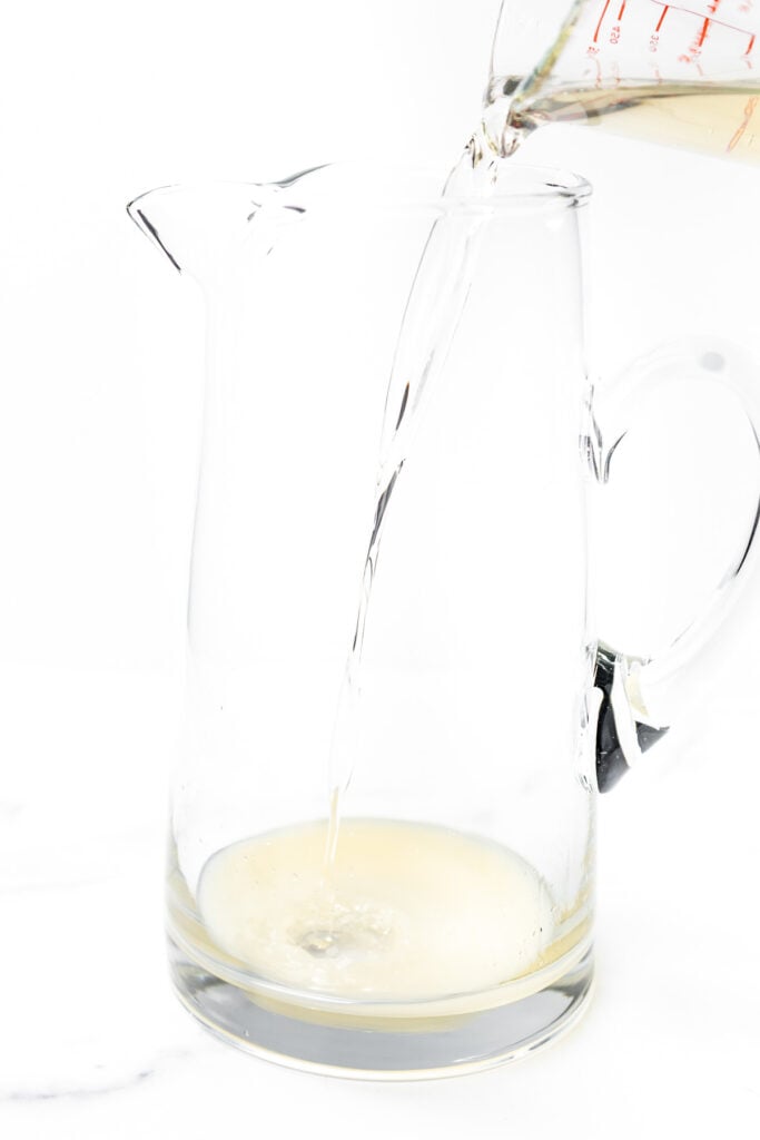 A clear liquid is being poured from a measuring cup into a glass pitcher containing a small amount of light-colored liquid.